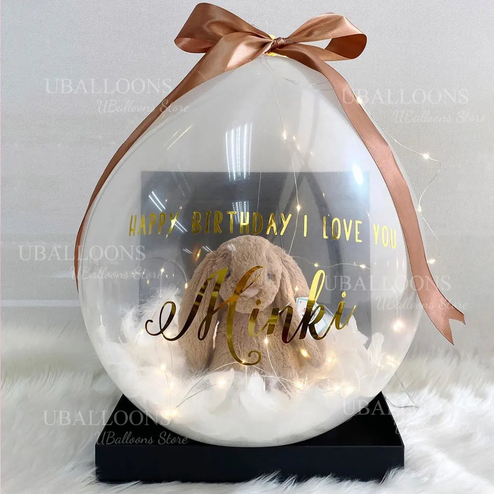 A clear latex balloon filled with white fluff features a gold birthday message and is tied with a satin ribbon.