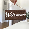 A rustic wooden welcome sign with white script 'Welcome' is being held by a hand, displayed on a white surface.