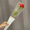 A hand holds a clear plastic tube containing a handmade crochet tulip bouquet with a red flower and green leaves.