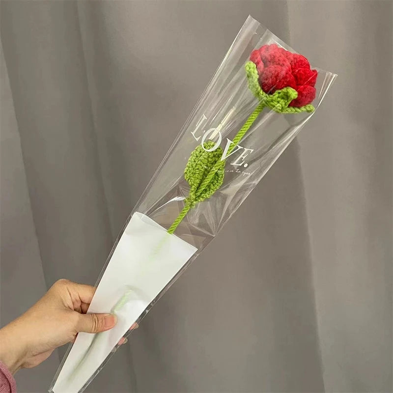 A hand holds a clear plastic tube containing a handmade crochet tulip bouquet with a red flower and green leaves.