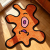 An orange cartoon monster shaped rug with purple eyes and black outline lying on a light wood floor.