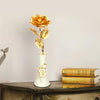 A golden metallic foil rose flower displayed in a white vase on a dark wooden surface.