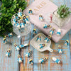 A collection of mini Virgin Mary resin statues in blue and white are displayed on a wooden surface next to a pink book.