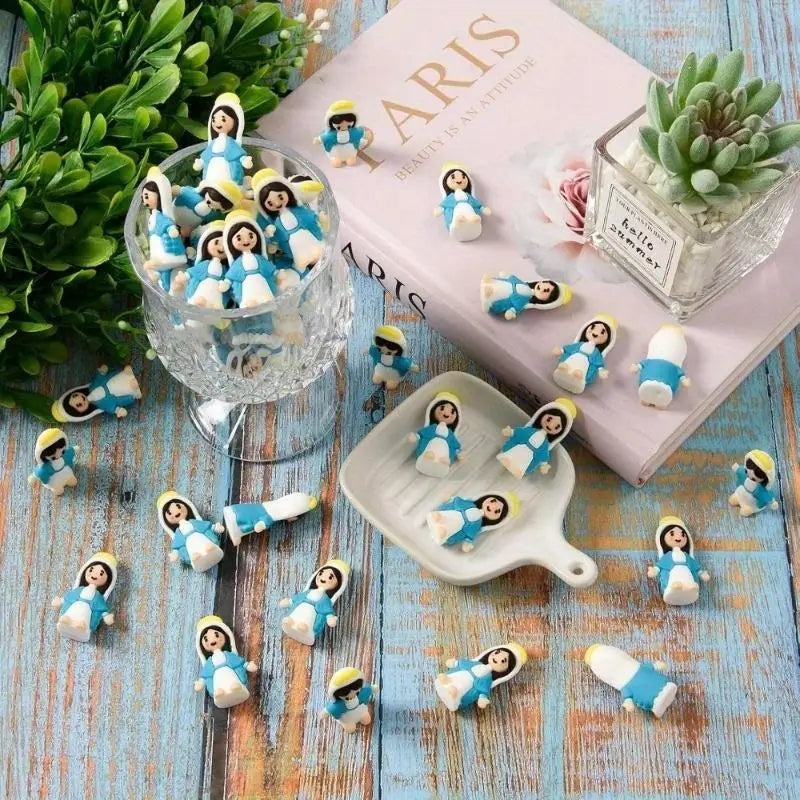 A collection of mini Virgin Mary resin statues in blue and white are displayed on a wooden surface next to a pink book.