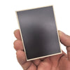 A hand holds a rectangular dark gray fridge magnet with light wood trim, featuring a ghost or pumpkin design.