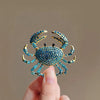 A hand holds a vintage marine life brooch pin set featuring a blue textured crab design.