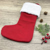 A red knitted Christmas alphabet sock with a white fluffy trim lying on a wooden surface next to pine cones and greenery.