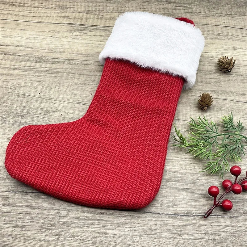 A red knitted Christmas alphabet sock with a white fluffy trim lying on a wooden surface next to pine cones and greenery.