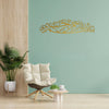 Gold Arabic calligraphy wall art featuring the Bismillah written on a light teal wall with a potted plant and an armchair.