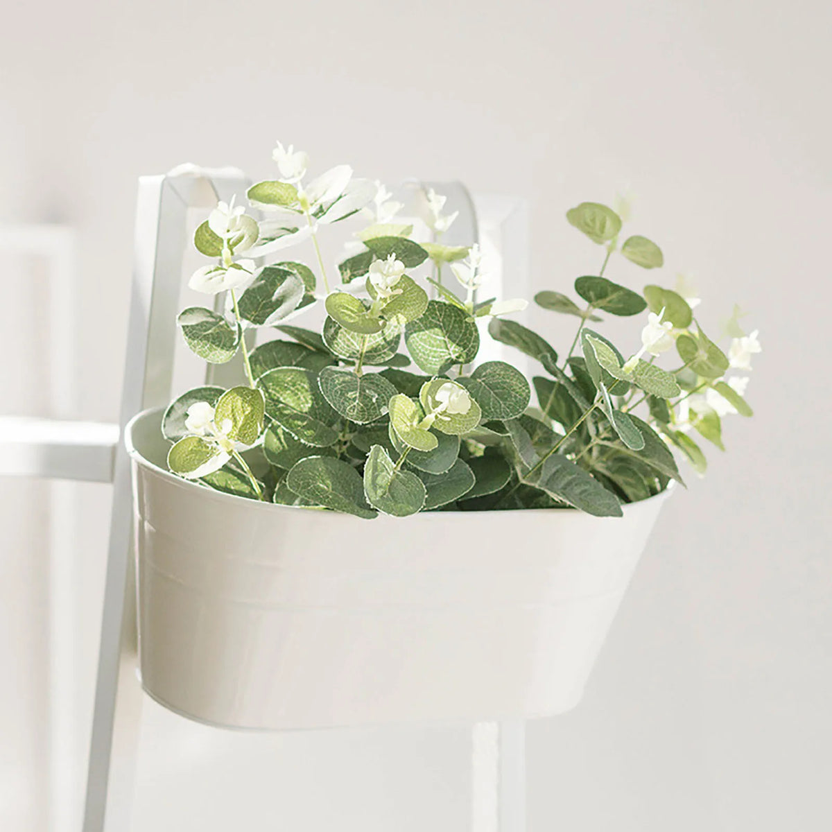 A white planter filled with green foliage and small white flowers is hanging on a white metal stand.