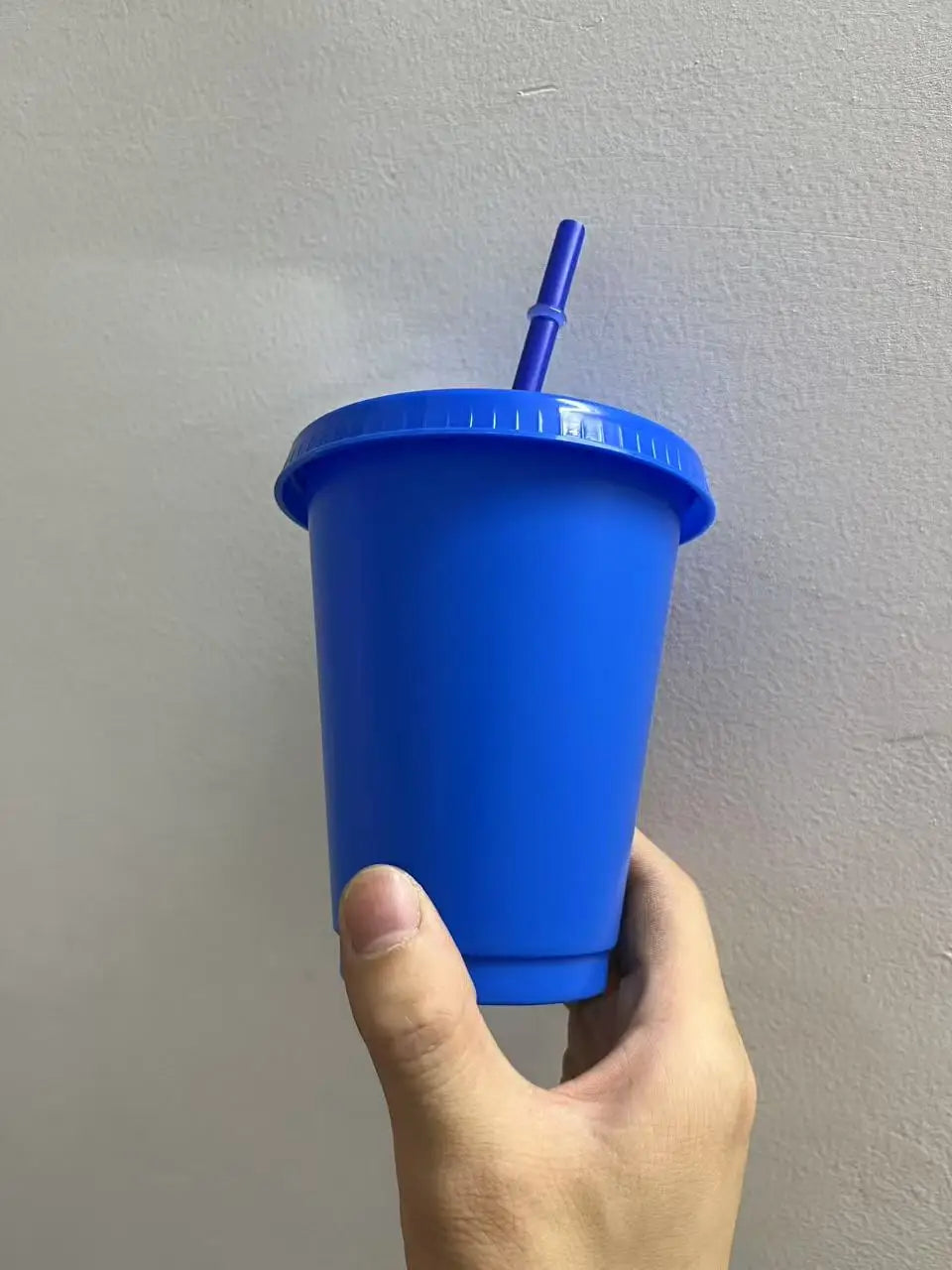 A hand holds a bright blue plastic cold cup with a lid and straw against a light gray wall.