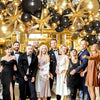 A group of people posing in front of a lavish black and gold balloon garland arch decoration.