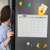 A close-up of a magnetic fridge planner with a monthly grid and colorful sticky notes on a dark gray refrigerator.