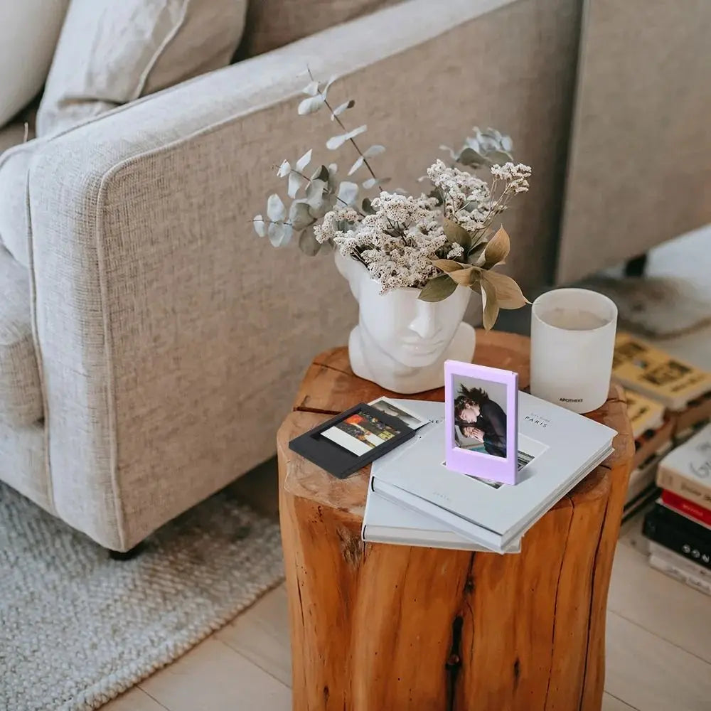 A small white ceramic vase holds dried greenery and white flowers on a wooden table next to a mini film frame.