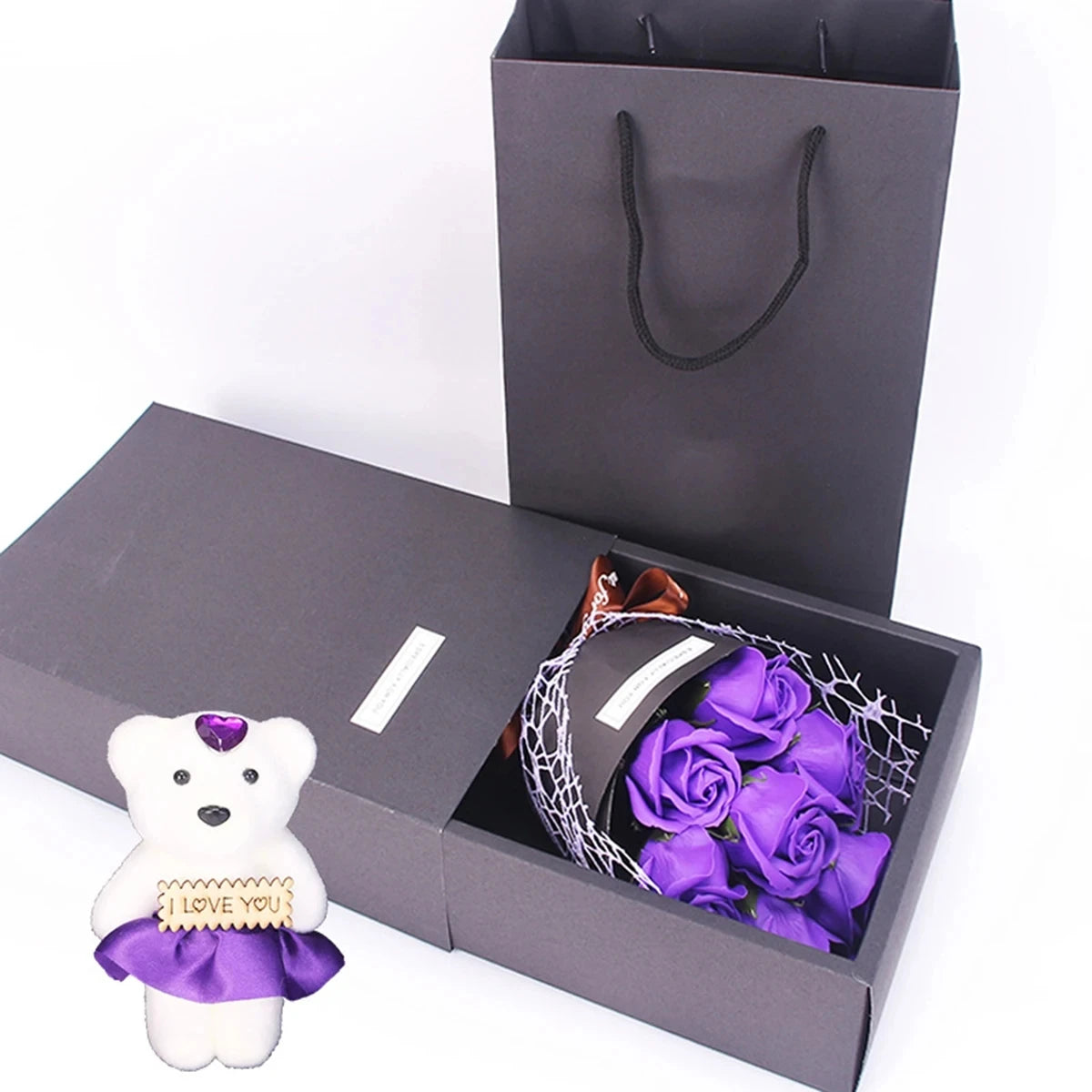 Una caja de regalo gris oscuro con un ramo de rosas moradas y una figura de oso blanco con una cinta de 'te amo'.