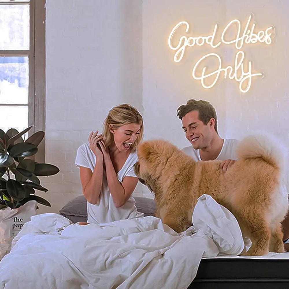 A blonde woman laughs while petting a fluffy golden dog in a bright room under a neon sign that reads Good Vibes Only
