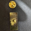 Gold enamel lapel pin featuring a chocolate factory ticket design on a dark surface.