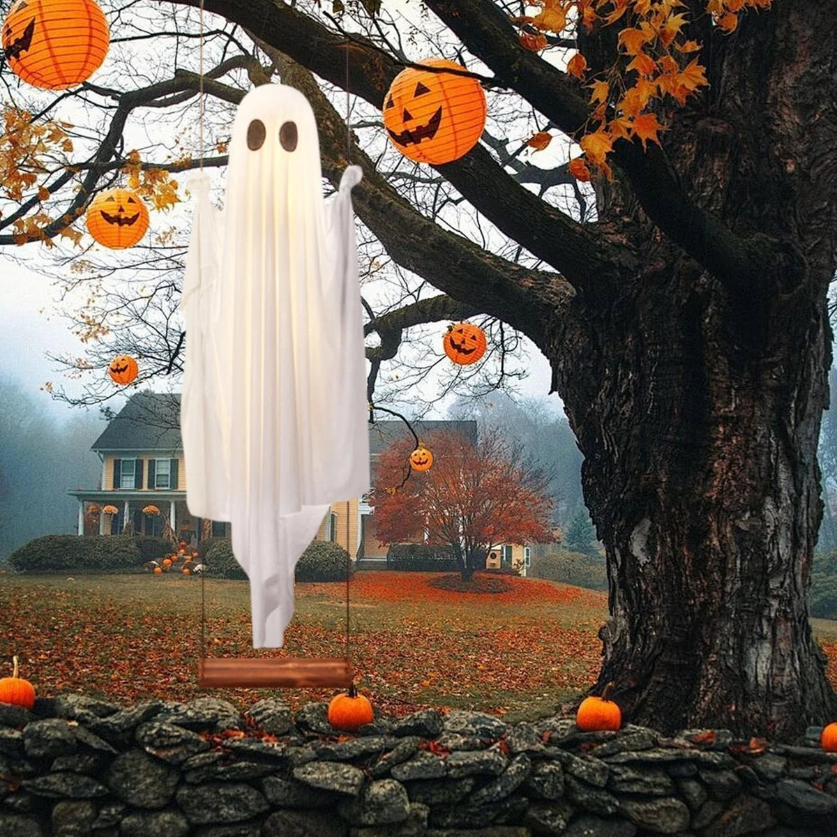 A white ghost decoration hangs from a tree branch in an outdoor autumn setting with orange pumpkins visible.