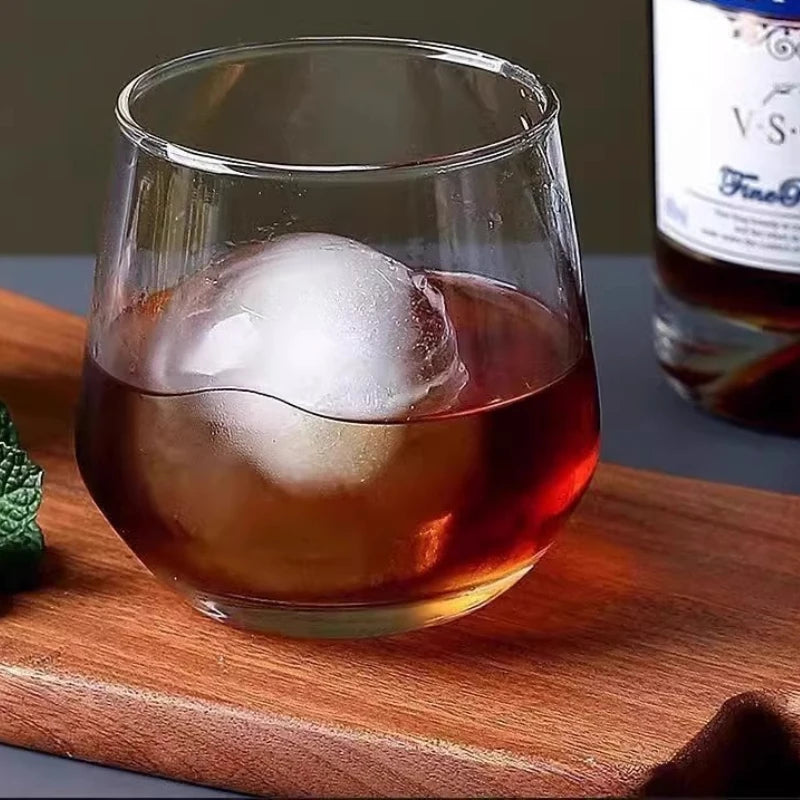 A glass of amber liquid with a large spherical ice cube resting on a wooden cutting board.