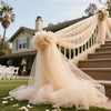 A sheer beige tulle fabric with a ruffled neckline and long drape is displayed on a grassy lawn next to stone stairs.