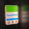 A rectangular white sign for FamilyMart with a green top section and blue stripe, featuring icons for coffee, cutlery, and 24