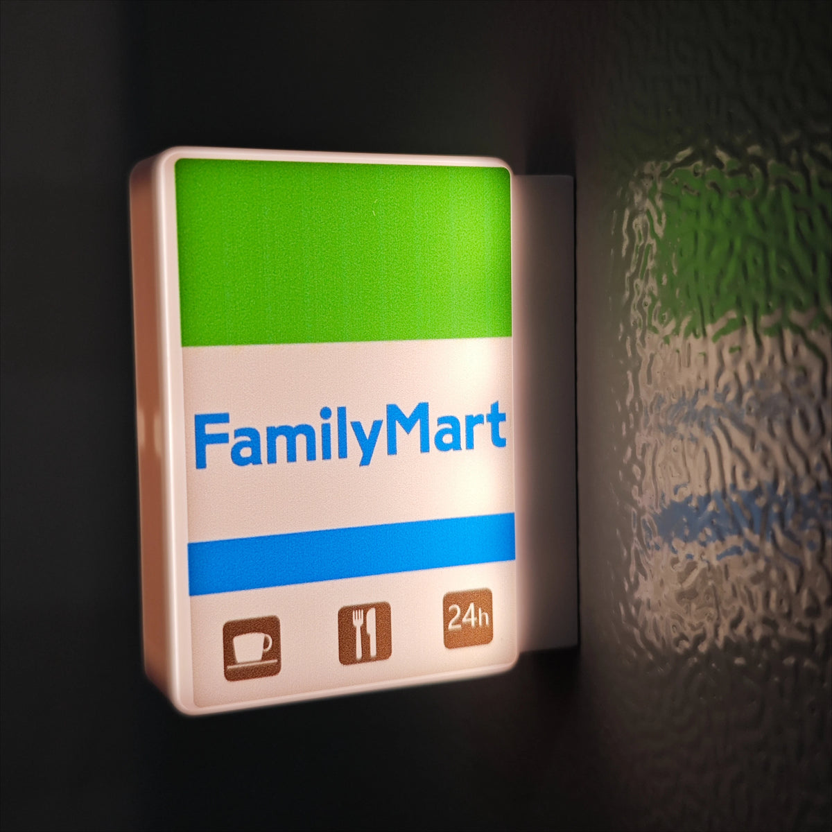 A rectangular white sign for FamilyMart with a green top section and blue stripe, featuring icons for coffee, cutlery, and 24