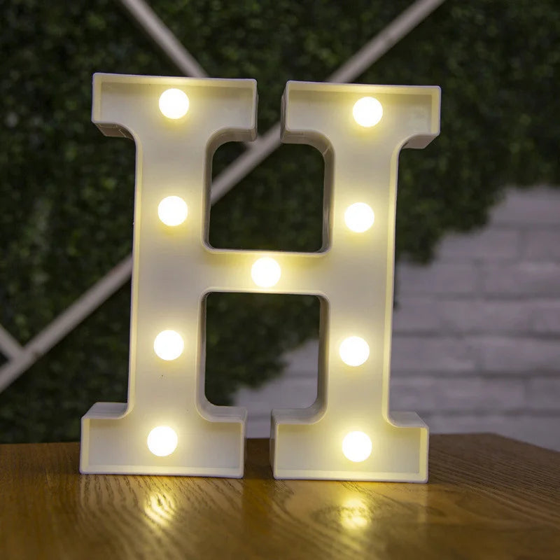 A glowing warm white LED alphabet letter light shaped like the letter H resting on a wooden surface.