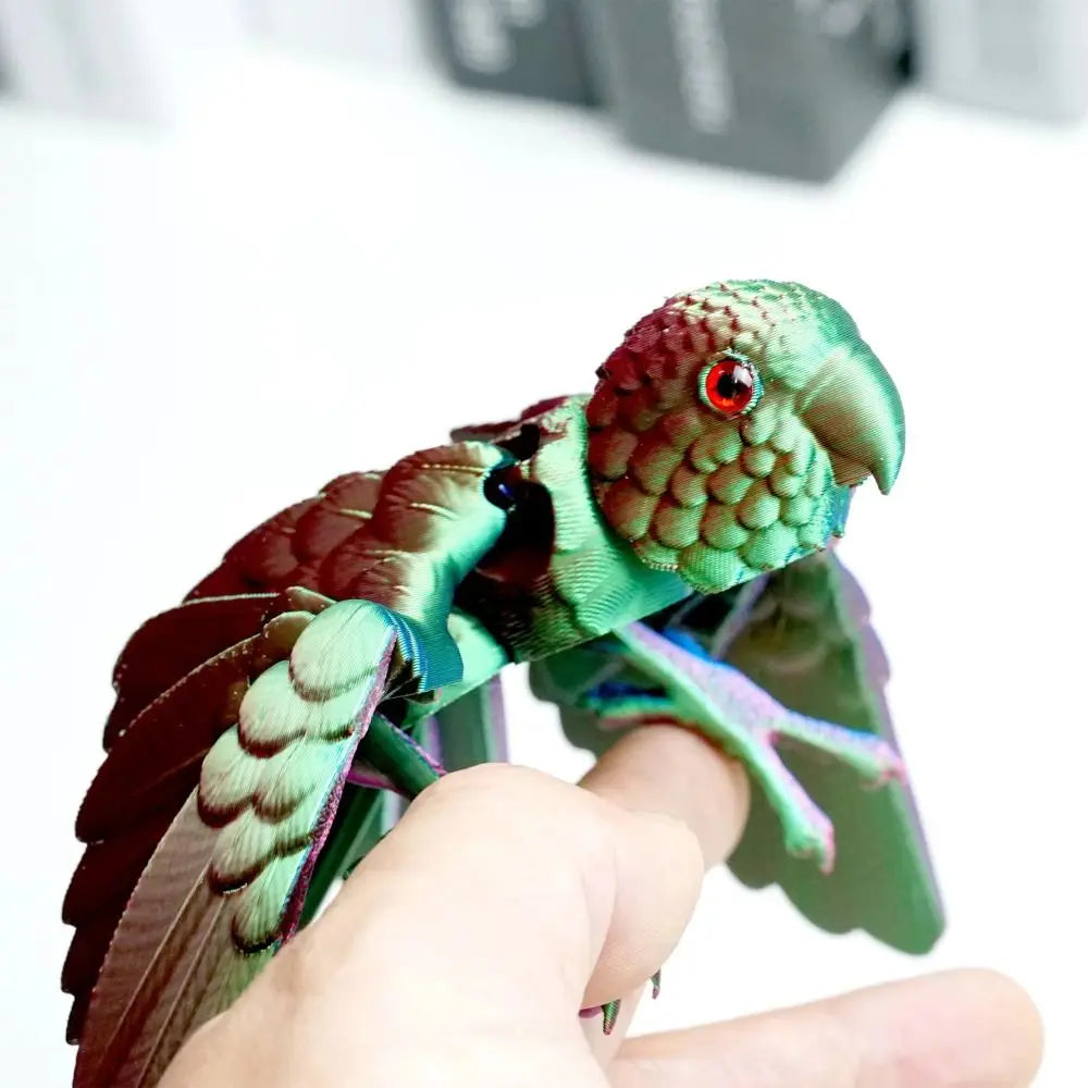 A hand holds an articulated 3D printed parrot figurine with movable wings, featuring green and reddish-brown plumage.
