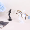 A minimalist black glass holder displaying a pair of round clear eyeglasses on a light pink surface.