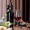 A dark glass bottle of red wine stands next to a stemmed glass, decorated with black satin ribbon bows and surrounded by flor