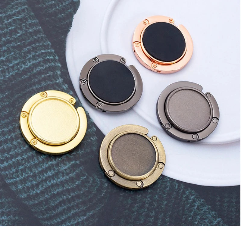 Four compact folding table bag hooks in gold, rose gold, black, and bronze tones arranged on a white plate.