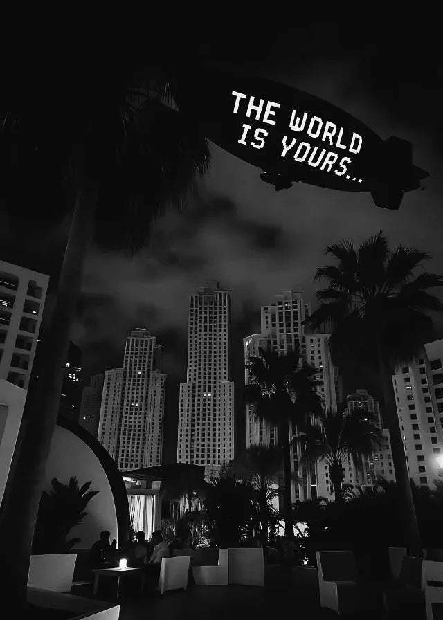 Black and white night photo of a palm tree silhouetted against a city skyline with a sign reading The World Is Yours
