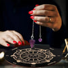 A hand with red nail polish holds a silver chain pendant over a wooden divination board featuring a sun symbol.