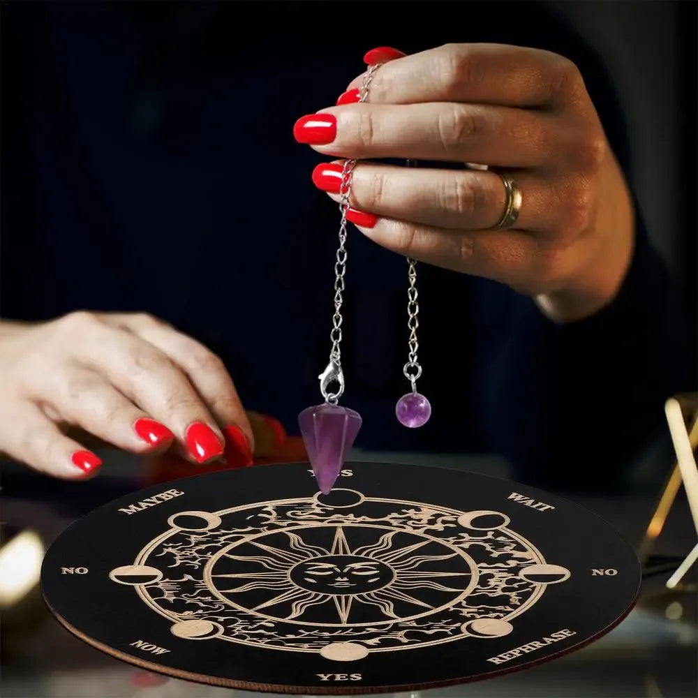 A hand with red nail polish holds a silver chain pendant over a wooden divination board featuring a sun symbol.