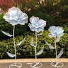 A set of three large, light blue velvet mesh flowers on slender stems displayed against a green hedge background.