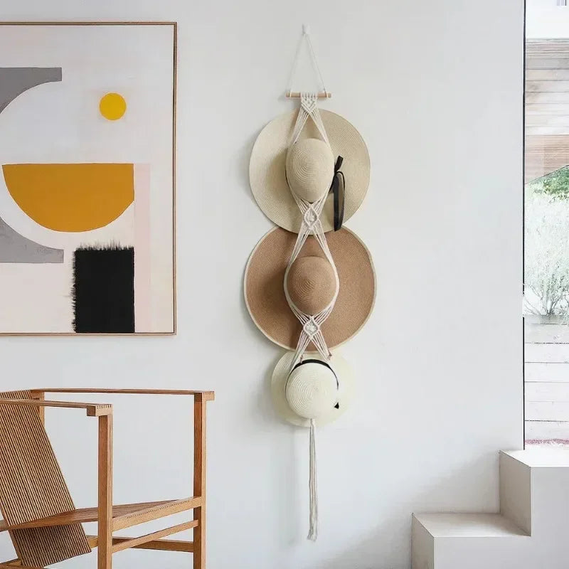 A handmade macrame hat organizer with a wooden dowel is displayed as a boho wall hanging on a white wall.