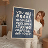 A woman holds up a framed canvas print with the text You are brave resilient precious strong confident and loved on it.