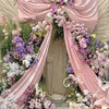 A floral archway decorated with pink satin draping and purple and white flowers for an event backdrop.