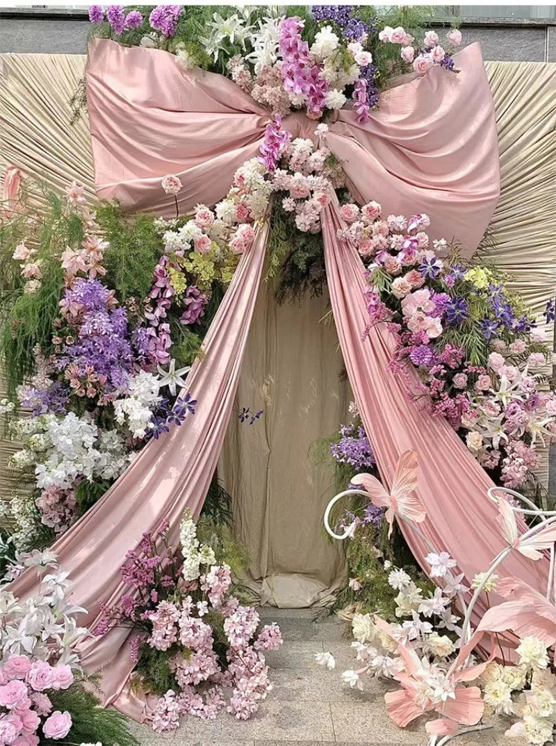 A floral archway decorated with pink satin draping and purple and white flowers for an event backdrop.