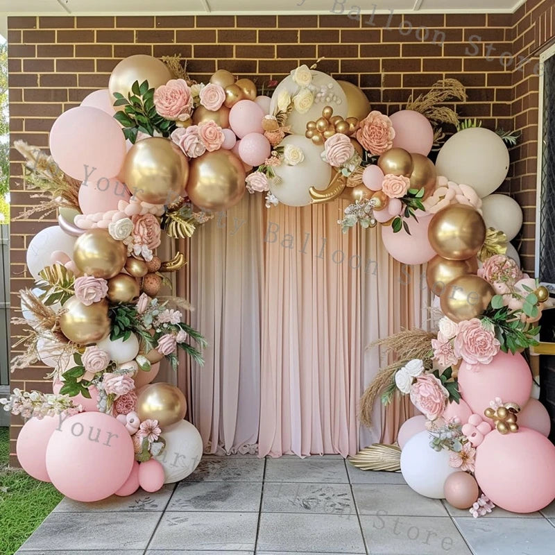A balloon garland arch featuring pink and gold balloons and floral greenery against a brick wall backdrop.