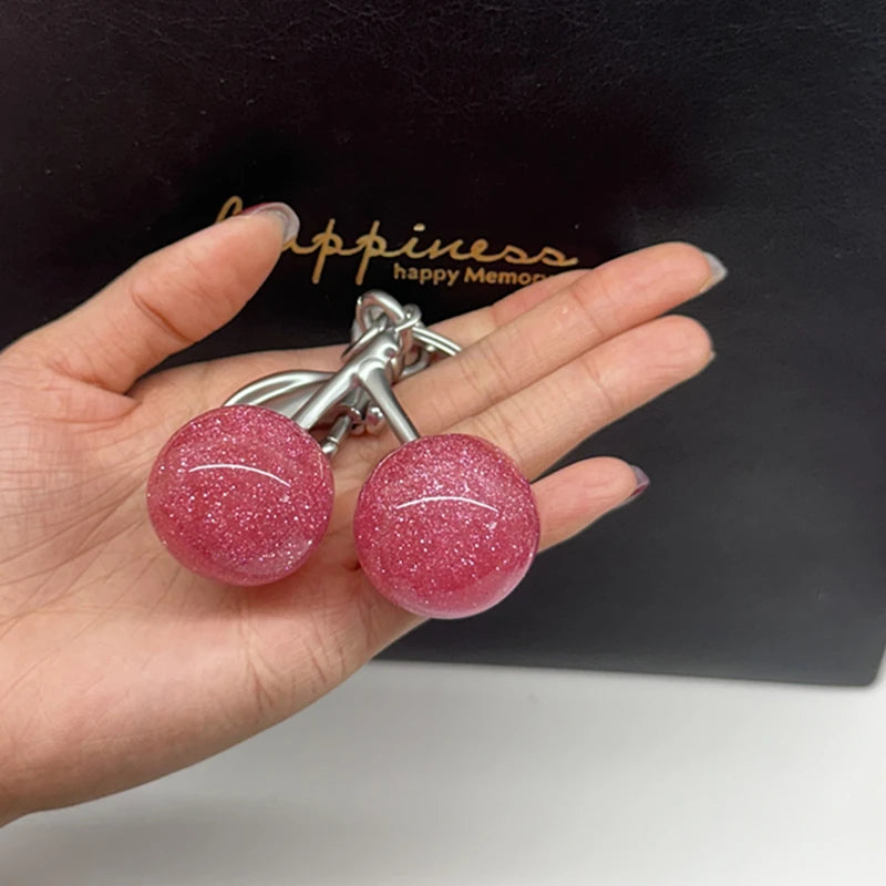 A hand holds a pink glittery cherry charm bag pendant with a silver keychain on a black background.