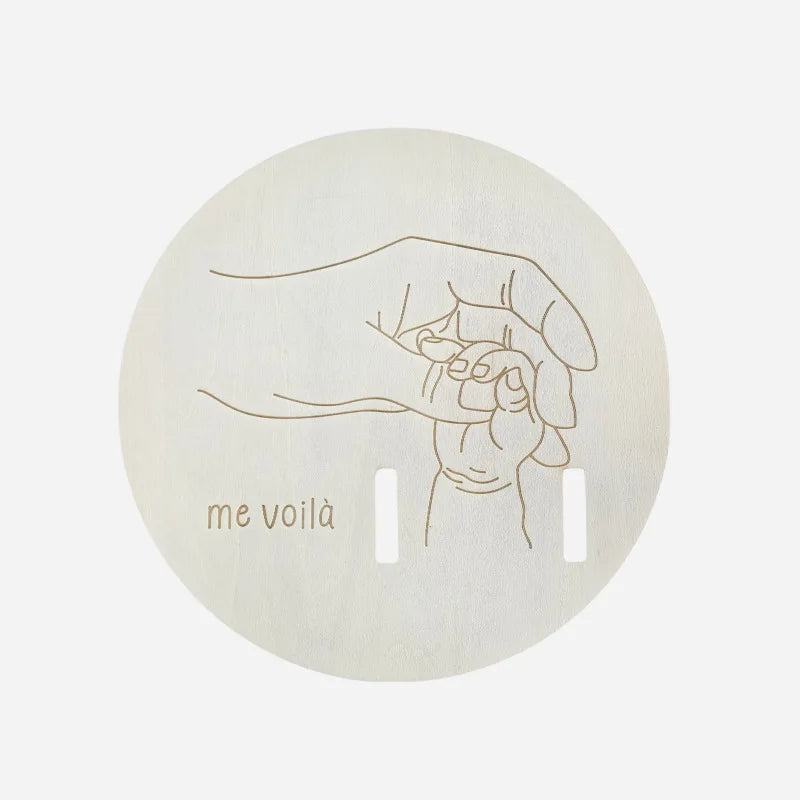 A light wood disc featuring a line drawing of a hand holding a finger, with the text me voilà.