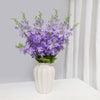 Un bouquet de fleurs artificielles de delphinium violet dans un vase blanc sur une surface blanche.