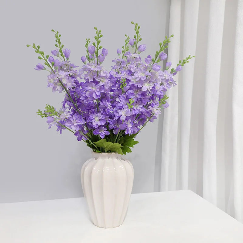 Un bouquet de fleurs artificielles de delphinium violet dans un vase blanc sur une surface blanche.