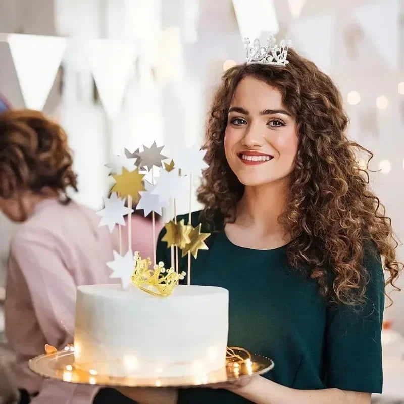 A young woman holds a white cake decorated with gold and silver crown toppers in a bright indoor setting.