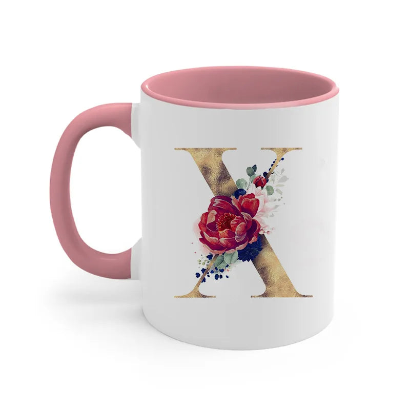A pink ceramic coffee mug featuring a gold letter X decorated with floral accents.