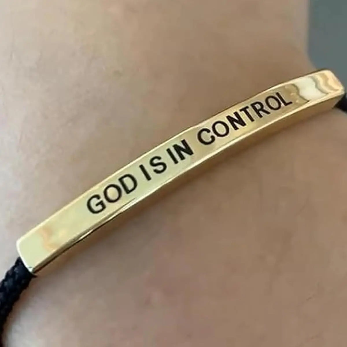 A gold-toned stainless steel bracelet with the inscription God Is in Control is shown against a skin background.