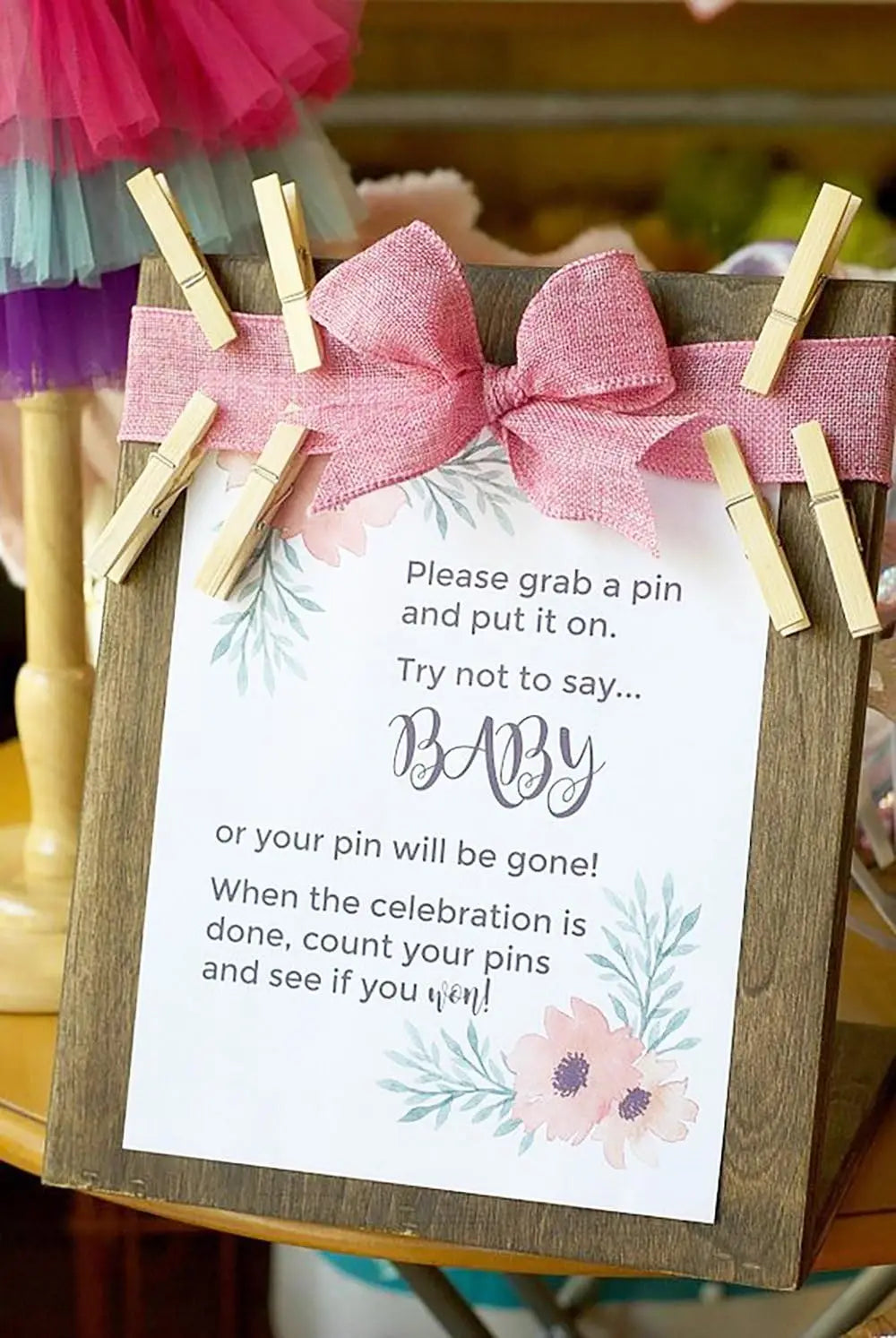 A wooden sign with a pink ribbon and text instructing guests to grab a pin for a celebration.