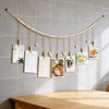 A beige beaded garland with wooden clips displays several framed photos and cards on a gray tiled wall.