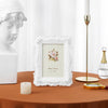 A white ornate photo frame with a pink rose design sits on a brown table next to a white statue and a gold candle holder.